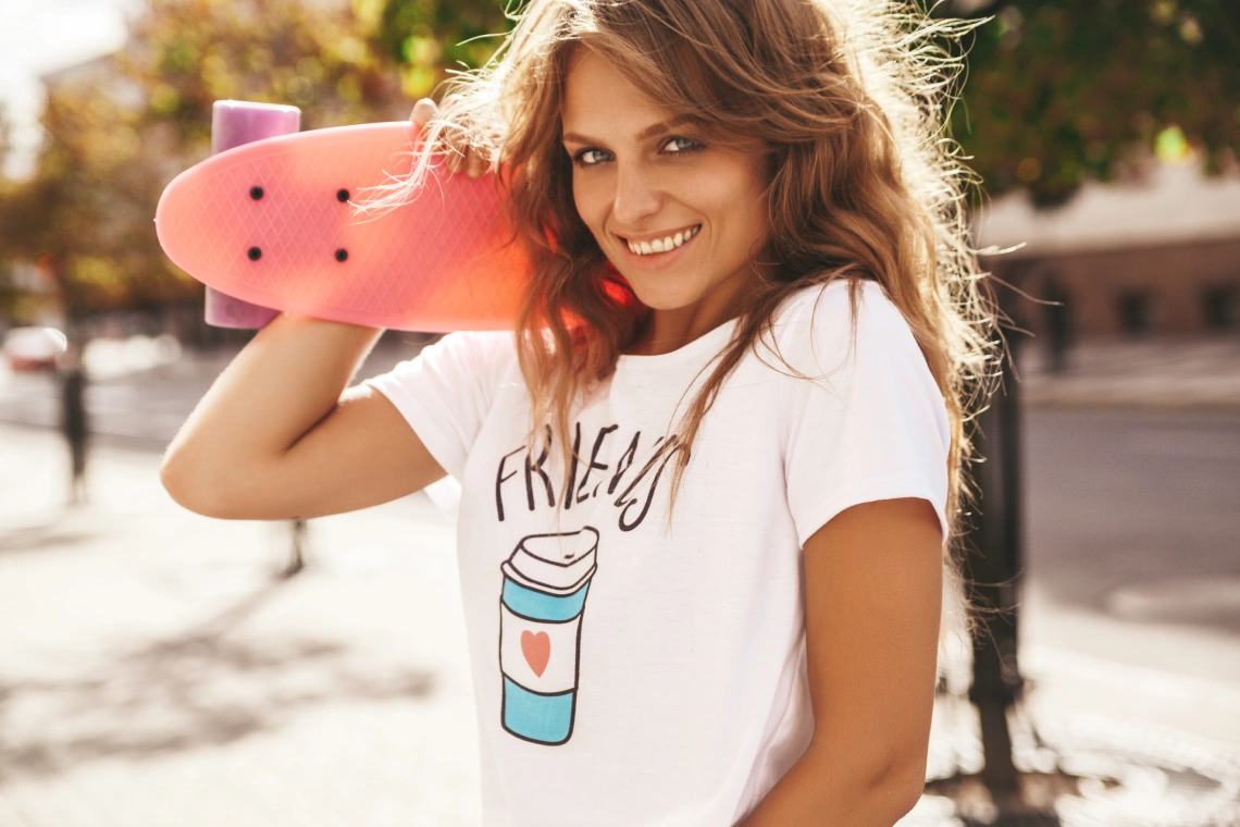 hermosa modelo de adolescente rubia linda y sonriente sin maquillaje en ropa blanca hipster de verano con patineta rosa centavo posando en el fondo de la calle